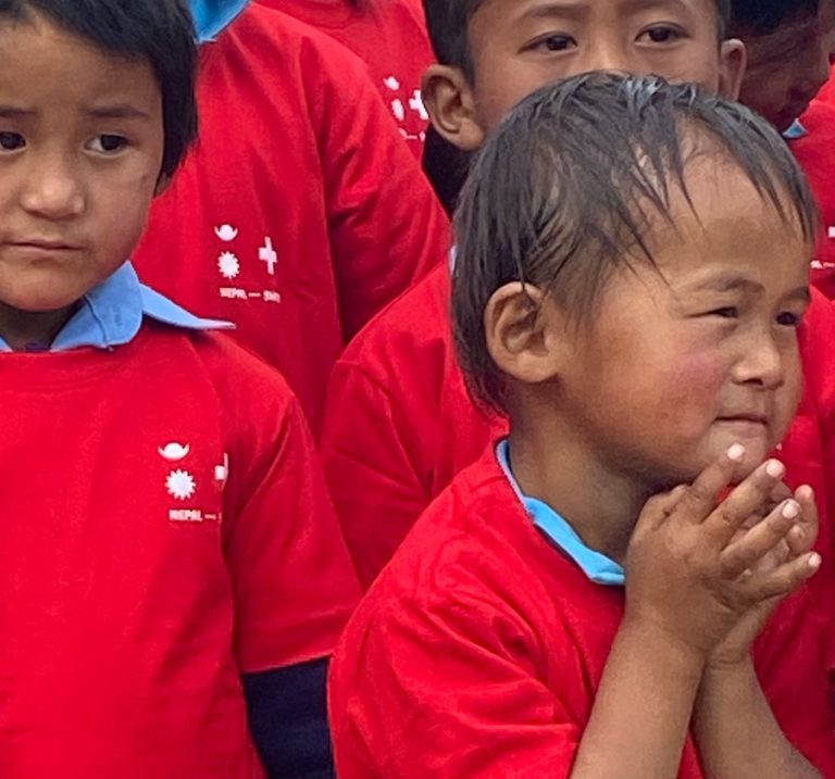 Zwei Kinder in roten Nepal-Swiss-Shirts, eines hält die Hände vor dem Gesicht zusammen.