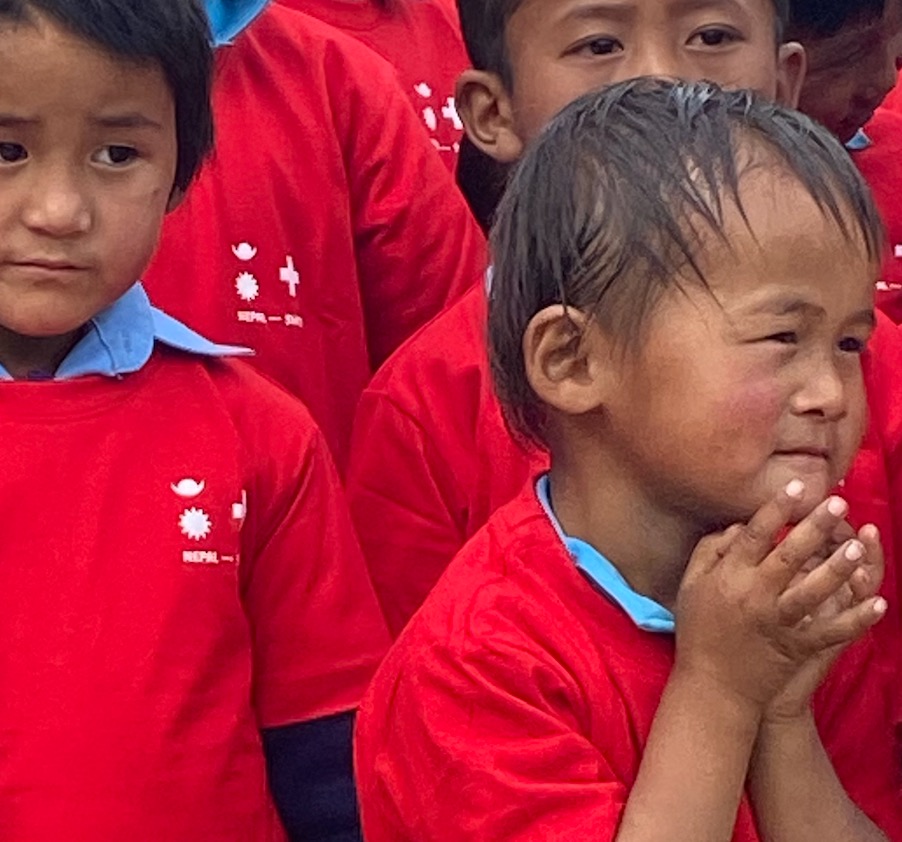 Zwei Kinder in roten Nepal-Swiss-Shirts, eines hält die Hände vor dem Gesicht zusammen.