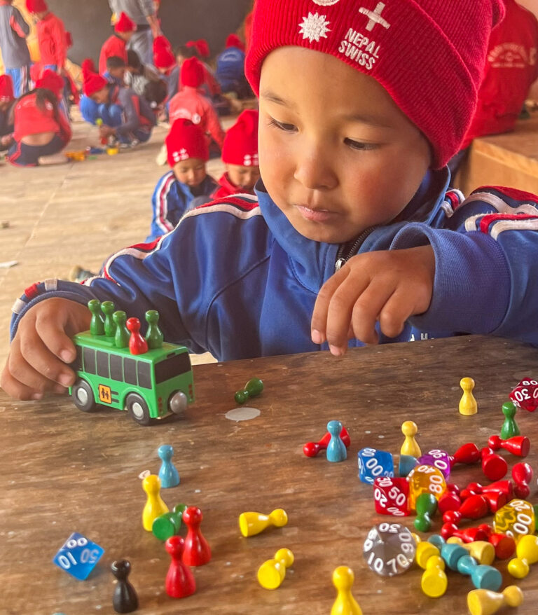 Ein Kind mit roter Nepal-Swiss-Mütze schiebt einen grünen Spielzeugbus über einen Tisch voller Spielsteine und Würfel.