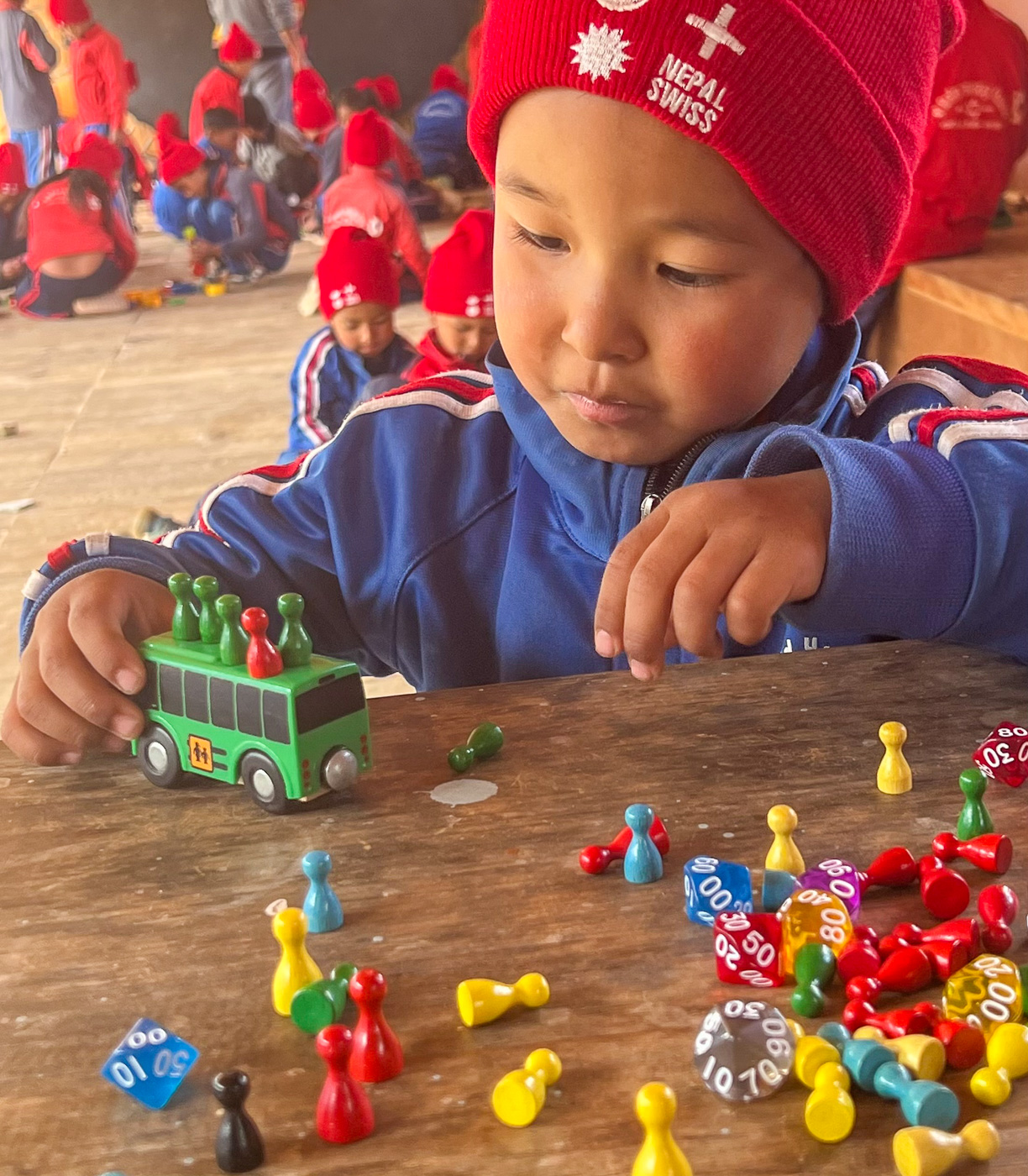 Ein Kind mit roter Nepal-Swiss-Mütze schiebt einen grünen Spielzeugbus über einen Tisch voller Spielsteine und Würfel.