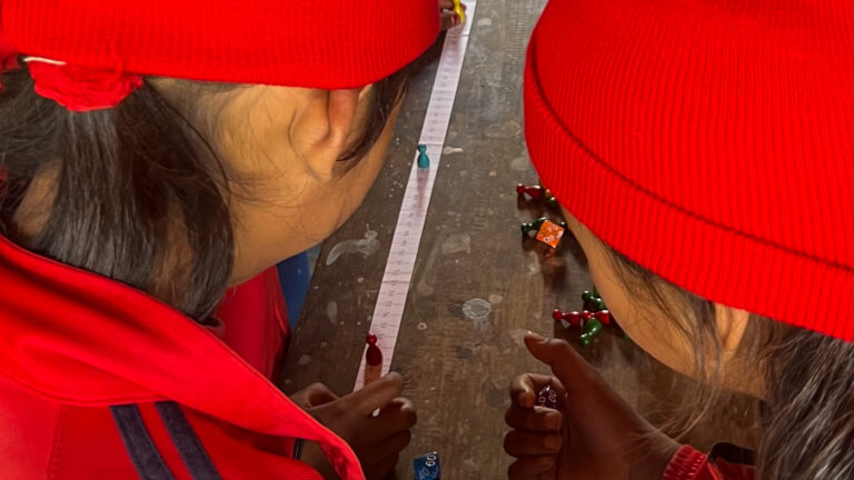 Zwei Schulkinder mit roten Mützen messen am Boden mit einem Massband; daneben liegen bunte Spielsteine.