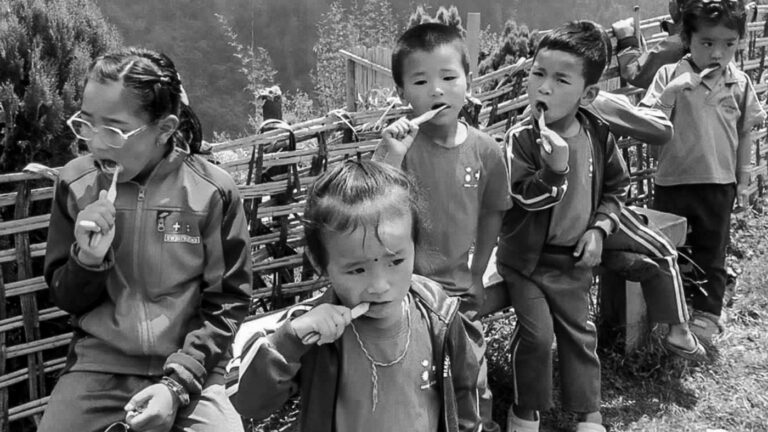 Fünf Kinder sitzen an einem Flechtzaun und putzen sich die Zähne. Schwarzweiss.